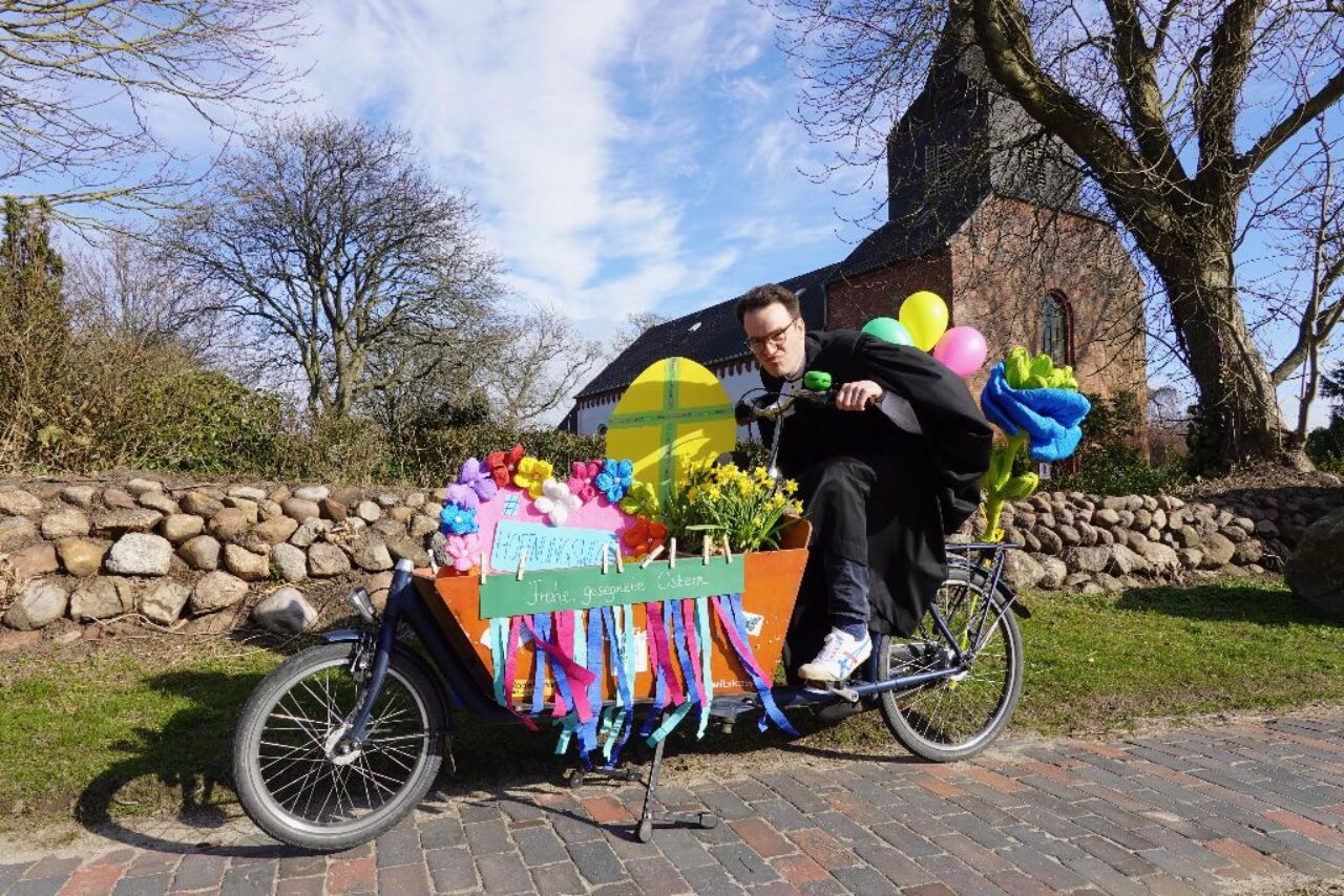 Pastor im Talar auf österlich geschmücktem Lastenfahrrad